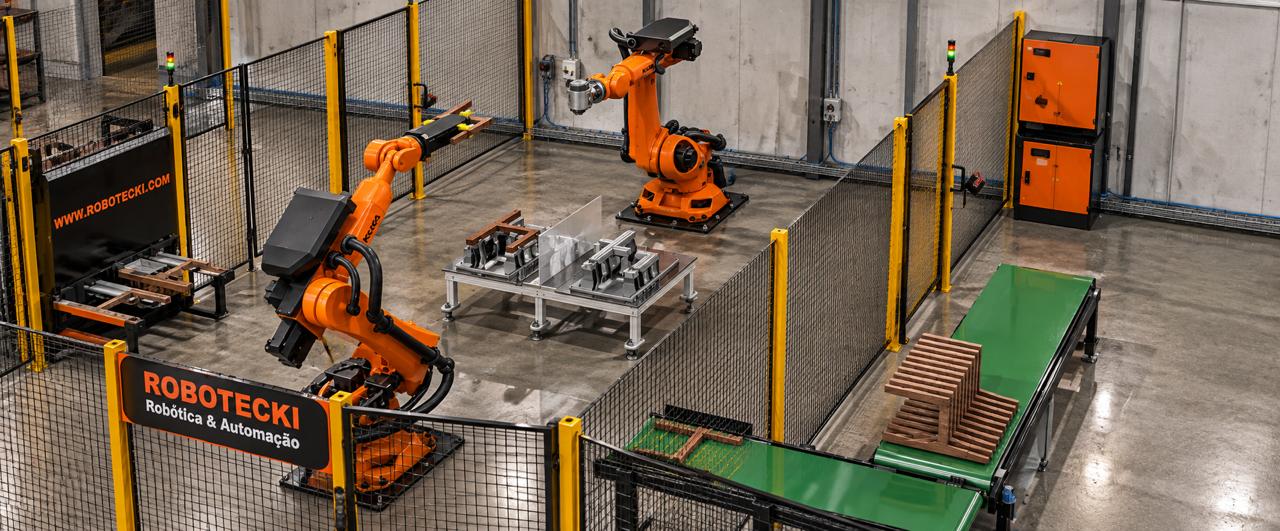 Célula Robotecki com dois robôs industriais, dispositivos de fixação, esteiras e cercamento de proteção